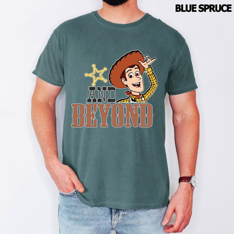 May include: Blue Spruce t-shirt with a cartoon image of Woody from Toy Story. The design includes a sheriff's badge, the words "AND BEYOND" in brown, and Woody. The shirt is a solid colour and has short sleeves.