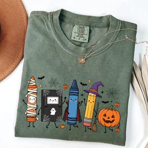 Pode incluir: Uma t-shirt verde-oliva com uma estampa gráfica de um giz de cera múmia, um livro preto com um fantasma, um giz de cera vampiro azul, um lápis bruxa amarelo e uma abóbora. O design inclui pequenos morcegos, teias de aranha e abóboras.