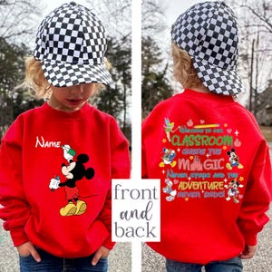 Puede incluir: Una sudadera roja con Mickey Mouse sosteniendo un bol&iacute;grafo, con la palabra "Name" arriba. La parte posterior de la sudadera tiene el texto "Welcome to our classroom where the magic never stops and the adventure never ends" con personajes de Disney.