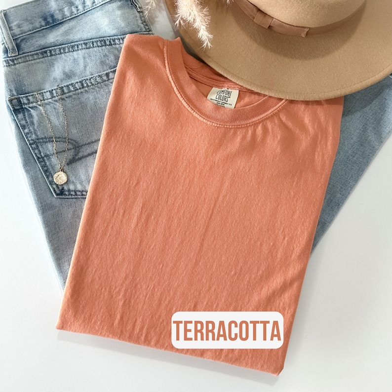 Puede incluir: Una camiseta de cuello redondo en color terracota liso con una etiqueta de la marca Comfort Colors en el interior del cuello. La prenda se coloca sobre unos vaqueros de mezclilla azul claro, junto a un sombrero beige de ala ancha y un collar dorado.