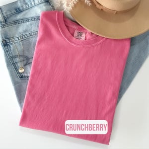 Puede incluir: Una camiseta de cuello redondo en color rosa s&oacute;lido con una etiqueta Comfort Colors en el interior del cuello y el texto CRUNCHBERRY impreso en rosa sobre un fondo rectangular blanco. La prenda se coloca sobre vaqueros azul claro y un sombrero.