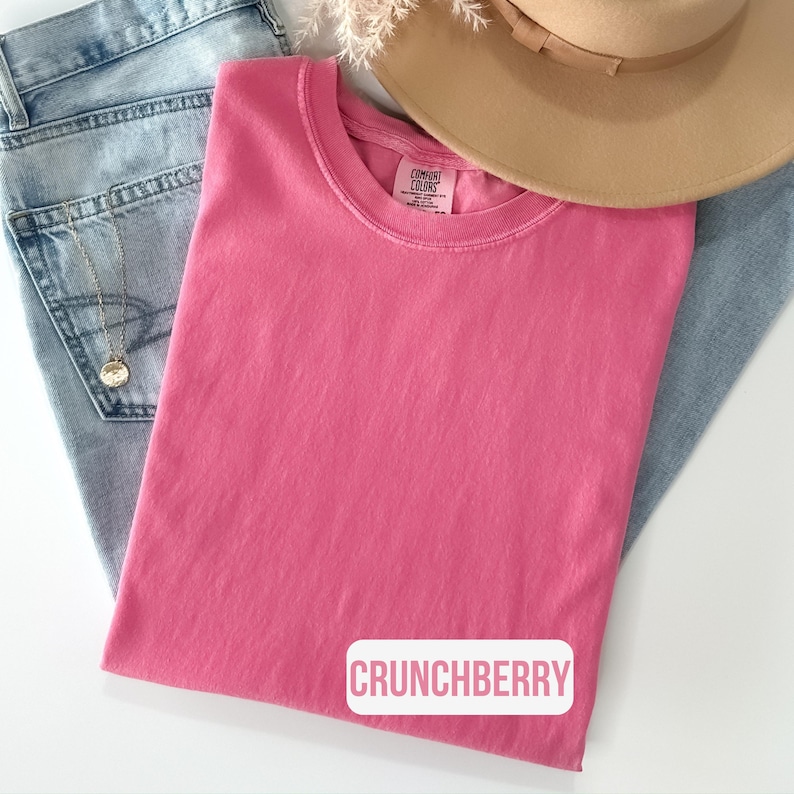 Puede incluir: Una camiseta de cuello redondo en color rosa s&oacute;lido con una etiqueta Comfort Colors en el interior del cuello y el texto CRUNCHBERRY impreso en rosa sobre un fondo rectangular blanco. La prenda se coloca sobre vaqueros azul claro y un sombrero.