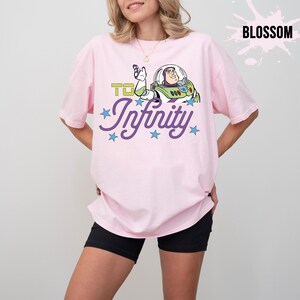 May include: A pale pink t-shirt with a Buzz Lightyear graphic and the words "To Infinity" in green and "Infinity" in purple script, with blue stars. The shirt has a relaxed, oversized fit.