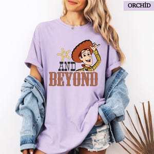 May include: Orchid purple t-shirt with a cartoon image of Woody from Toy Story. The design includes a sheriff's badge, Woody's hat, and the words "AND BEYOND" in brown. The shirt has short sleeves and a loose fit.