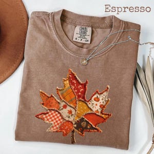 May include: An espresso-coloured short-sleeve t-shirt featuring a large, central graphic of a maple leaf composed of various patchwork fabric patterns, including orange plaid, red floral, and pumpkin prints, with a raw-edge frayed appliqué style.