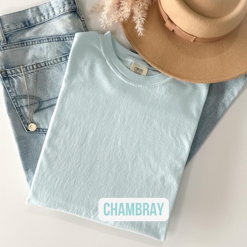 Puede incluir: Una camiseta de cuello redondo azul claro con etiqueta Comfort Colors, junto a unos vaqueros azul claro, un sombrero beige de ala ancha y una delicada cadena dorada con un colgante circular. El texto CHAMBRAY es visible.