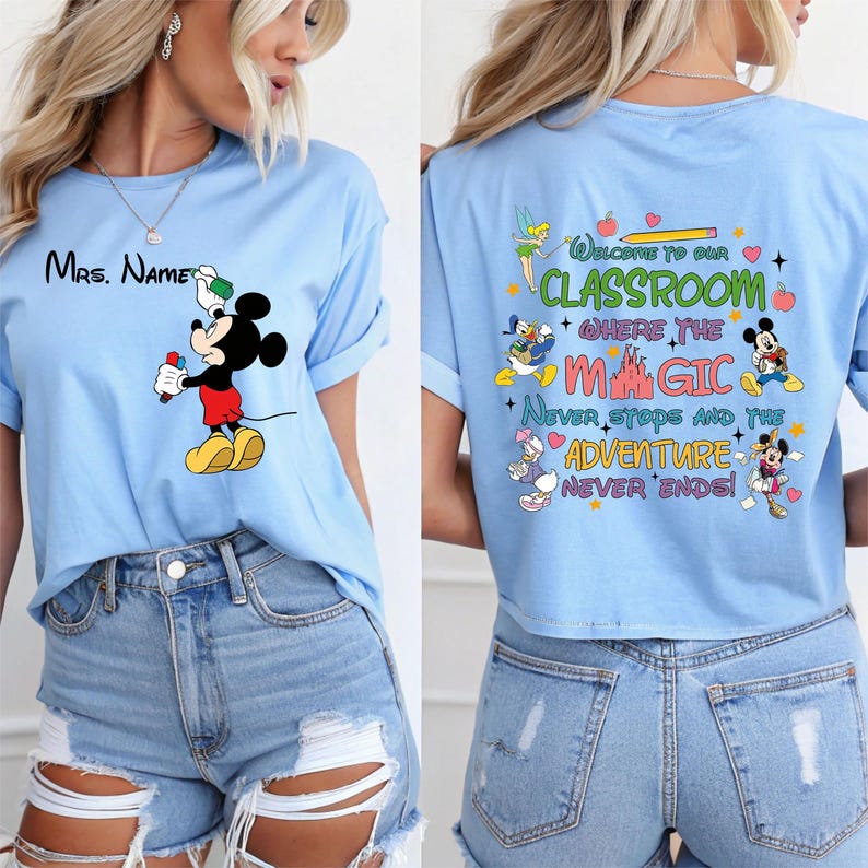 Puede incluir: Camiseta azul claro con Mickey Mouse sosteniendo una bebida y el texto "Mrs. Name" en la parte delantera. La parte trasera de la camiseta tiene el texto "Welcome to our Classroom where the Magic Never Stops and the Adventure Never Ends!" con personajes de Disney.