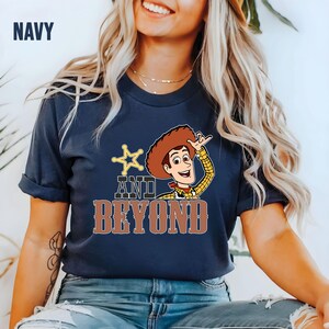 May include: Navy blue t-shirt with a cartoon image of Woody from Toy Story. The shirt features the text "AND BEYOND" in brown letters, with a star and Woody's image. The word "NAVY" is in the upper left corner.