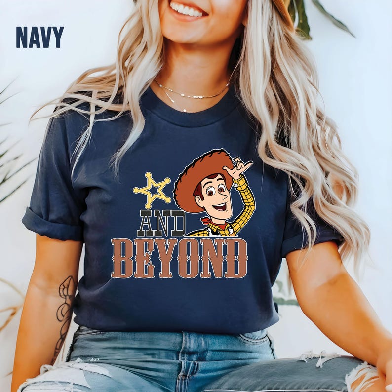 May include: Navy blue t-shirt with a cartoon image of Woody from Toy Story. The shirt features the text "AND BEYOND" in brown letters, with a star and Woody's image. The word "NAVY" is in the upper left corner.