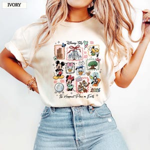 May include: Ivory-colored t-shirt featuring a colorful Disney Trip design. The design includes various Disney characters and landmarks, with the text "2026" and "The Happiest Place on Earth".