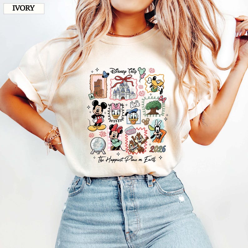 May include: Ivory-colored t-shirt featuring a colorful Disney Trip design. The design includes various Disney characters and landmarks, with the text "2026" and "The Happiest Place on Earth".