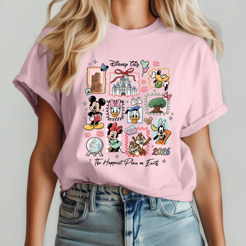 May include: A light pink t-shirt featuring a colorful graphic design with Disney characters and landmarks. The design includes Mickey Mouse, Minnie Mouse, Donald Duck, and Chip and Dale. Text on the shirt reads "Disney Trip" and "The Happiest Place on Earth" with the year "2026".