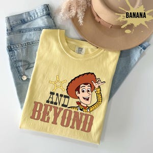 May include: A yellow t-shirt with a cartoon Woody design and the words "AND BEYOND". The image includes light blue jeans and a tan cowboy hat. The word "BANANA" is printed on the hat. The shirt is a comfortable fit.