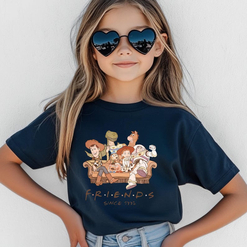 Pu&ograve; includere: T-shirt blu navy con una grafica dei personaggi di Toy Story seduti su un divano. La parola "FRIENDS" &egrave; stampata sotto la grafica, con "SINCE 1995" sotto. La persona nell'immagine indossa occhiali da sole a forma di cuore.