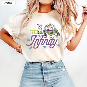 May include: Ivory t-shirt with a Buzz Lightyear graphic and the words "To Infinity". The design features green and purple text with blue star details. The shirt is a casual style with a crew neck.
