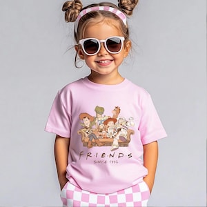 Pu&ograve; includere: Una bambina indossa una maglietta rosa con un disegno di Toy Story, con la scritta "FRIENDS SINCE 1995". La bambina indossa anche occhiali da sole e una fascia rosa e bianca.
