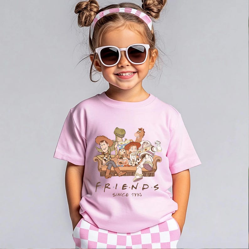 Pu&ograve; includere: Una bambina indossa una maglietta rosa con un disegno di Toy Story, con la scritta "FRIENDS SINCE 1995". La bambina indossa anche occhiali da sole e una fascia rosa e bianca.