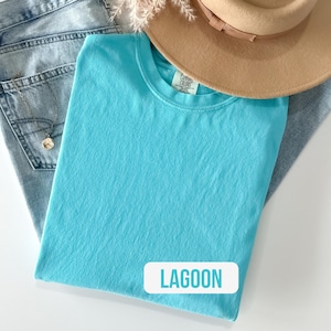 Puede incluir: Una camiseta de cuello redondo en color turquesa brillante con una etiqueta de Comfort Colors en el interior del cuello. La prenda se coloca sobre unos vaqueros azul claro y bajo un sombrero beige de ala ancha. Se ve un collar dorado con un colgante.