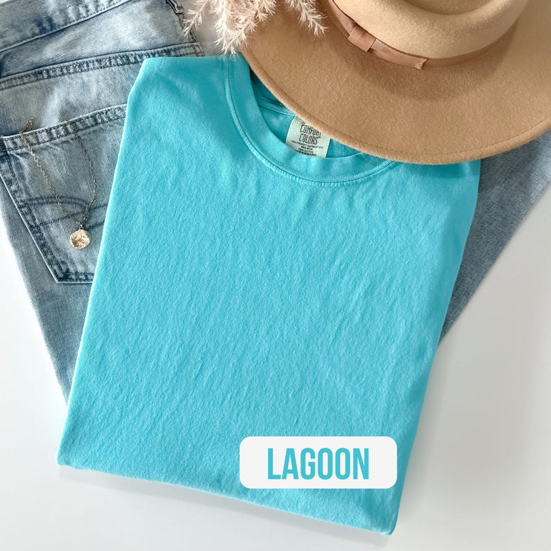 Puede incluir: Una camiseta de cuello redondo en color turquesa brillante con una etiqueta de Comfort Colors en el interior del cuello. La prenda se coloca sobre unos vaqueros azul claro y bajo un sombrero beige de ala ancha. Se ve un collar dorado con un colgante.