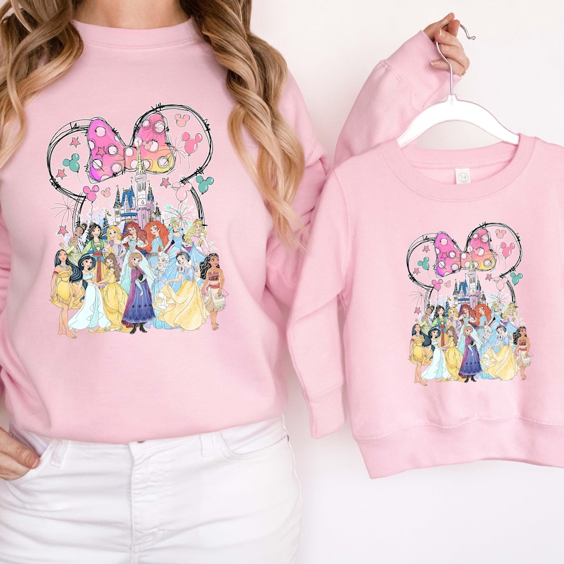 May include: Pink sweatshirts featuring a Minnie Mouse-inspired design with a bow and a collection of Disney princesses. The design includes a castle and fireworks. One sweatshirt is adult-sized, and the other is child-sized.