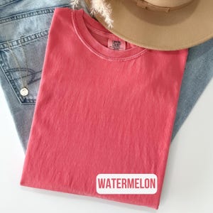 Könnte beinhalten: Ein einfarbiges, korallenrosa T-Shirt mit kurzen Ärmeln, Rundhalsausschnitt und einem Comfort Colors Markenetikett am Innenkragen. Das T-Shirt zeigt das Wort WATERMELON in weißen Großbuchstaben auf einem rechteckigen Hintergrund unten rechts.