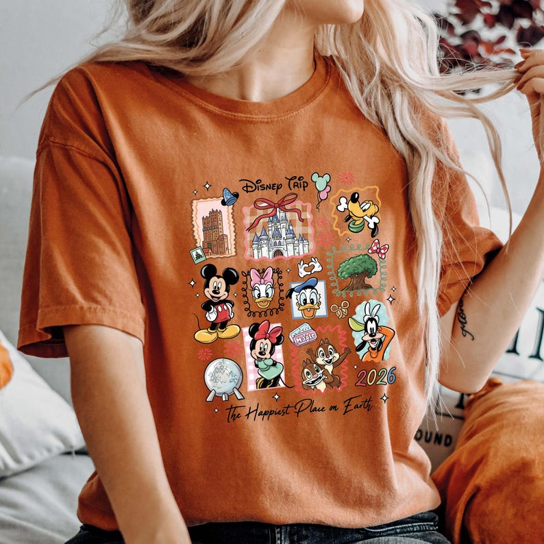 May include: Burnt orange t-shirt featuring a colorful Disney Trip graphic. The design includes various Disney characters, a castle, and the text "2026" and "The Happiest Place on Earth."