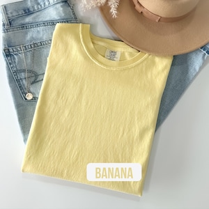 Puede incluir: Una camiseta de cuello redondo en color amarillo claro liso con una etiqueta de Comfort Colors en el interior del cuello. La prenda se coloca sobre unos vaqueros azul claro y se acompa&ntilde;a de un sombrero beige de ala ancha y un collar de plata.