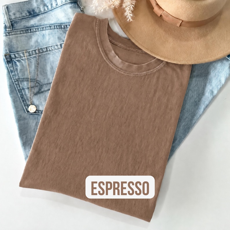 Puede incluir: Una camiseta de cuello redondo en color marr&oacute;n espresso con una etiqueta rectangular blanca que muestra el texto ESPRESSO impreso en marr&oacute;n. La prenda se presenta sobre unos vaqueros azul claro, un sombrero beige y un collar de cadena plateada.