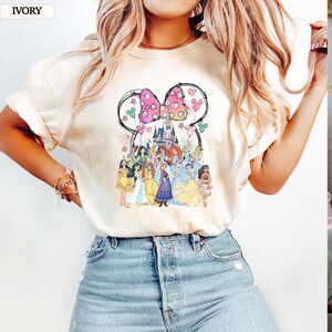 May include: Ivory-colored t-shirt featuring a colorful graphic of Disney princesses and a castle. The design includes a Minnie Mouse bow and Mickey Mouse silhouettes. The word "IVORY" is printed in the top left corner.