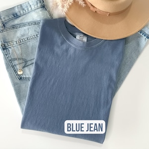 Puede incluir: Una camiseta azul lisa de cuello redondo con etiqueta Comfort Colors, colocada junto a unos vaqueros de lavado claro, un sombrero beige de ala ancha y un collar con colgante dorado. El texto BLUE JEAN aparece en un recuadro blanco abajo a la derecha.