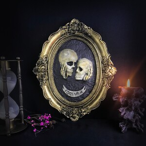 May include: A gold oval frame with a black background and two white skull sculptures. The skulls are facing each other and have a banner that reads "FOREVER".