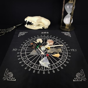 May include: A black cloth with a white circle and numbers printed on it. The circle has letters around the outside and a triangle in the center. There are various small objects on the cloth, including bones, a crystal, a skull, and a die.