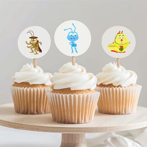 Puede incluir: Tres cupcakes con glaseado blanco, cada uno coronado con un adorno de papel circular sobre un palillo de madera. Los adornos presentan ilustraciones de dibujos animados de un saltamontes marrón, una hormiga azul y una oruga amarilla.
