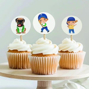 May include: Three cupcakes with white frosting topped with round paper circles. The circles feature illustrations of a boy in a baseball uniform, a boy in a blue shirt and a dog wearing a green shirt.
