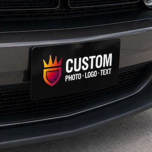 May include: A dark gray Dodge Challenger R/T front end featuring a black rectangular license plate with a colorful crown logo and the white text CUSTOM PHOTO LOGO TEXT. The vehicle has a prominent hood scoop and circular headlights.