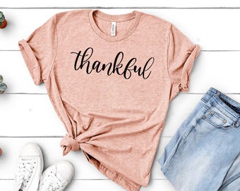 Puede incluir: Una camiseta de color melocotón con la palabra "thankful" escrita en letras cursivas negras.