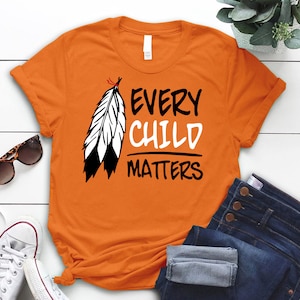 May include: Orange t-shirt with a black and white feather design and the text "Every Child Matters".