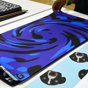 May include: A rectangular mousepad with a blue, black, and light blue swirl design. The mousepad is being held up, revealing a stack of other mousepads with various designs, including a leopard print and a bear face.