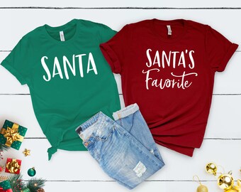 Può includere: Due magliette a tema natalizio. Una maglietta verde con la parola "SANTA" in lettere bianche. Una maglietta rossa con il testo "SANTA'S Favorite" in lettere bianche.