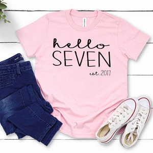 K&ouml;nnte beinhalten: Rosa T-Shirt mit dem Text "hello seven est. 2017" in schwarzer Schrift. Das Shirt liegt flach auf einer wei&szlig;en Holzoberfl&auml;che mit einer blauen Jeans und wei&szlig;en Turnschuhen.