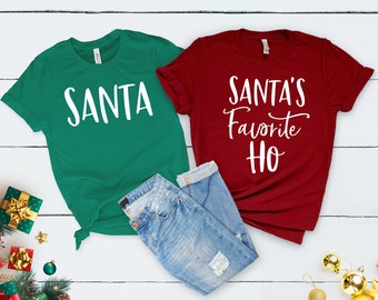 Può includere: Due magliette a tema natalizio. Una maglietta verde con la parola "SANTA" in lettere bianche. Una maglietta rossa con il testo "SANTA'S Favorite HO" in lettere bianche.