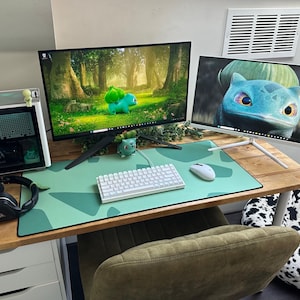 May include: A gaming setup featuring two computer monitors displaying images of a Bulbasaur. The desk includes a white keyboard, mouse, and a green mousepad. A computer tower, headphones, and a Bulbasaur figurine are also visible.