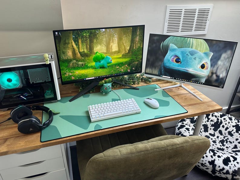 May include: A gaming setup featuring two computer monitors displaying images of a Bulbasaur. The desk includes a white keyboard, mouse, and a green mousepad. A computer tower, headphones, and a Bulbasaur figurine are also visible.