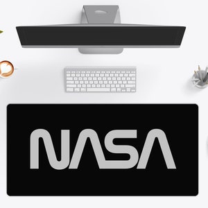Puede incluir: Alfombrilla de escritorio negra con el logotipo de la NASA en blanco. La alfombrilla rectangular está sobre un escritorio blanco, junto con un monitor de ordenador, teclado, ratón y una taza de café.