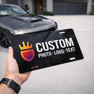 May include: Black license plate with the word "CUSTOM" in white, and the words "PHOTO • LOGO • TEXT" below. A colorful crown and shield logo is on the left. The plate is held in front of a black car.