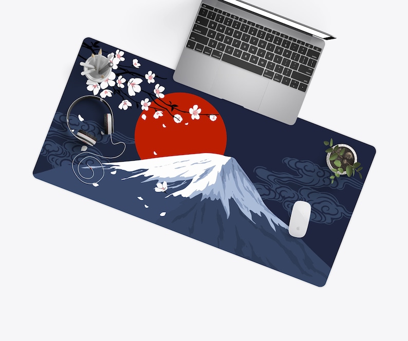May include: A navy blue desk mat featuring a Japanese-inspired design. The mat showcases a red sun, a snow-capped mountain, and cherry blossoms. A laptop, headphones, and a mouse are on the mat.