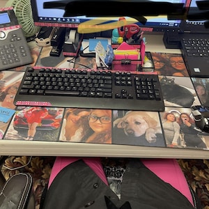 May include: A desk setup featuring a black keyboard with pink accents, a black mouse, and a phone. The desk has a custom mousepad with various photos. A laptop and monitor are also visible.