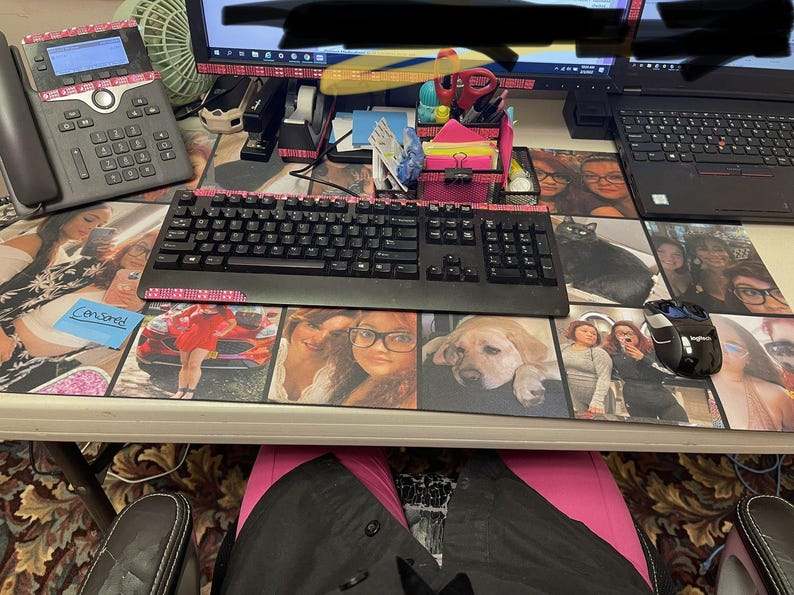 May include: A desk setup featuring a black keyboard with pink accents, a black mouse, and a phone. The desk has a custom mousepad with various photos. A laptop and monitor are also visible.