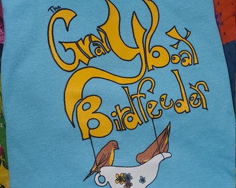 May include: A light blue t-shirt featuring a yellow graphic that reads "The Gray Box Birdfeeder" with two brown birds in a white teacup.
