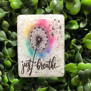 May include: A rectangular badge reel with a dandelion design. The design features a black dandelion with a watercolor background of pink, yellow, blue, and green. The words "just breathe" are written in black script. The reel has a glittery, round button.